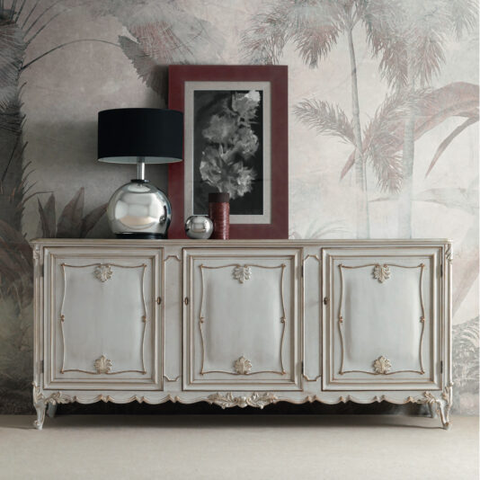 A Large Rococo Inspired Sideboard with intricate details is positioned against a wall adorned with tropical wallpaper. Atop the sideboard, there is a sleek modern silver lamp, a framed black-and-white artwork set against a burgundy mat, and a small vase. The entire arrangement seamlessly combines classic and contemporary elements.