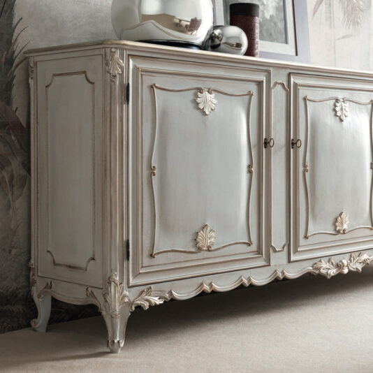 The Large Rococo Inspired Sideboard boasts intricate carvings and a distressed light gray finish, complete with decorative leaf accents and ornate legs. Two shiny silver decorative items adorn its top, creating an elegant contrast against the patterned wallpaper backdrop.