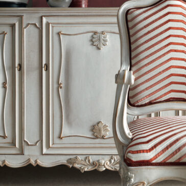 A close-up of a sophisticated vintage setting showcases a detailed white and gold cabinet adorned with intricate carvings, evoking the elegance of the Large Rococo Inspired Sideboard, paired with a wooden chair upholstered in red and white stripes. The decor exudes a timeless, luxurious appeal.