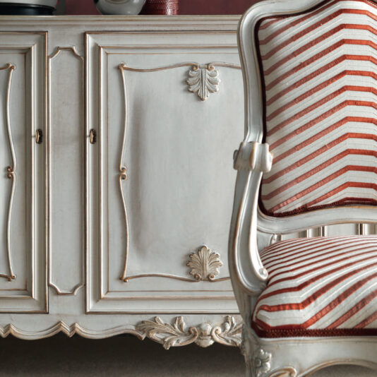 A close-up of a sophisticated vintage setting showcases a detailed white and gold cabinet adorned with intricate carvings, evoking the elegance of the Large Rococo Inspired Sideboard, paired with a wooden chair upholstered in red and white stripes. The decor exudes a timeless, luxurious appeal.