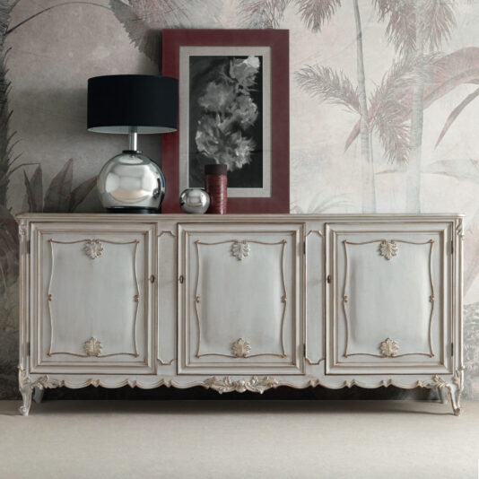 A Large Rococo Inspired Sideboard with intricate carvings is positioned against a wallpapered wall adorned with a palm motif. Atop the sideboard sits a metallic lamp featuring a black shade, alongside a red vase and an abstract art piece in a dark color palette framed beautifully.