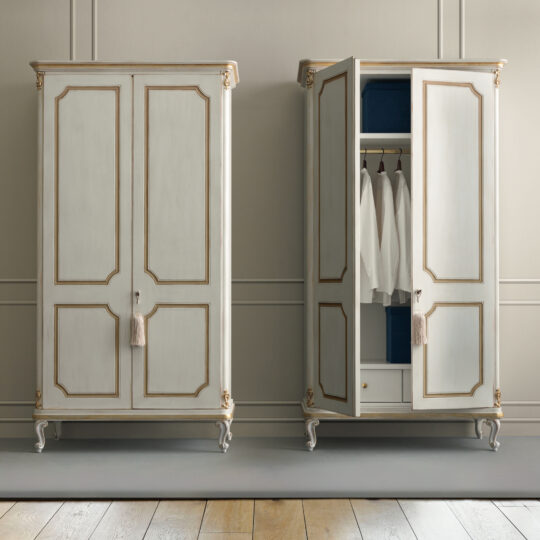 Against a light-colored wall stand two Narrow Classic Style Wardrobes featuring ornate detailing. One remains closed, while the other is open, showcasing white shirts hanging inside. The wooden floor further complements the elegant and timeless ambiance.