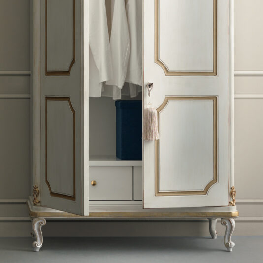 Narrow Classic Style Wardrobe A narrow, classic-style wardrobe in vintage cream with gold accents stands open, revealing white hanging clothes and a dark blue box on the bottom shelf. This wardrobe features ornate legs and a decorative tassel hanging from the handle.