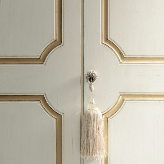 Narrow Classic Style Wardrobe Close-up of the Narrow Classic Style Wardrobe's ornate white and gold door, adorned with a decorative tassel hanging from the center. This large piece showcases carved geometric patterns and a textured appearance, enhancing its elegant design.