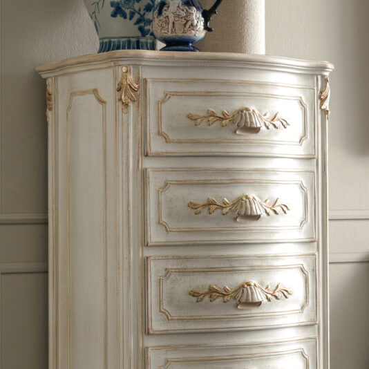 The Ornate Classic Style Tallboy, with its vintage-inspired design, features intricate gold detailing and a distressed white finish. Atop the tallboy, decorative porcelain vases—one boasting a blue floral pattern—enhance its charm. This timeless piece stands gracefully against a neutral-colored wall.