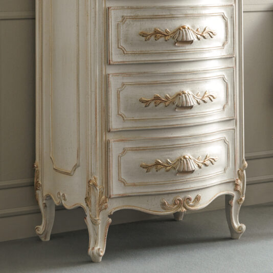 The Ornate Classic Style Tallboy features intricate carvings and three spacious drawers. It showcases a distressed white finish complemented by gold accents on the edges and decorative details. Resting gracefully on elegantly curved legs against a neutral-toned wall, this tallboy exudes timeless elegance.