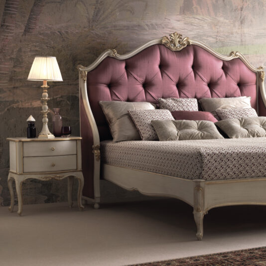 A vintage-style bedroom features a Classic Louis Style Button Upholstered Bed (Copy) with an ornate headboard in deep pink, complete with tufting. Decorative pillows and patterned sheets adorn the bed. Beside it is a wooden nightstand with a lamp, glass bottles, and a cup. The wallpaper has a subtle, elegant design.