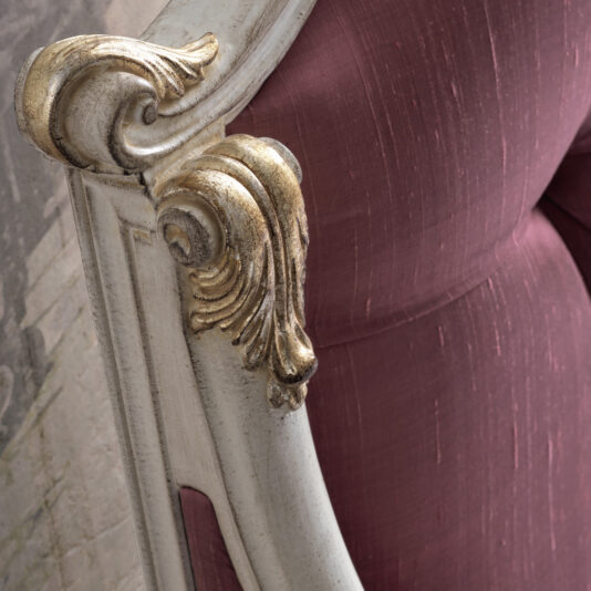 Close-up of an ornate cream-colored chair with gold accents on its carved wooden frame. The upholstery, inspired by the Classic Louis Style Button Upholstered Bed (Copy), is a deep mauve fabric. The intricate detailing and elegant design suggest a vintage or antique style.
