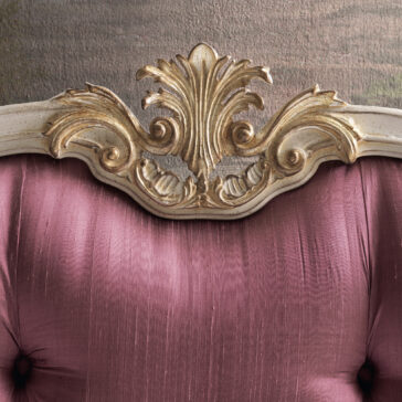 Close-up of the Classic Louis Style Button Upholstered Bed, showcasing a vintage-inspired headboard with a pink, tufted satin backrest reminiscent of an elegant Classic Louis design. The top edge is adorned with intricate gold and white wood carvings that highlight the detailed craftsmanship. The background has a soft, muted texture, creating a beautiful contrast that accentuates the bed's ornate details.