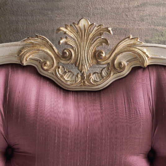 Close-up of the Classic Louis Style Button Upholstered Bed, showcasing a vintage-inspired headboard with a pink, tufted satin backrest reminiscent of an elegant Classic Louis design. The top edge is adorned with intricate gold and white wood carvings that highlight the detailed craftsmanship. The background has a soft, muted texture, creating a beautiful contrast that accentuates the bed's ornate details.