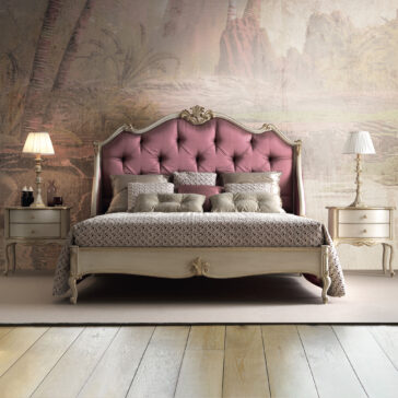 An elegant bedroom featuring a Classic Louis Style Button Upholstered Bed (Copy) with an ornate, tufted, pink headboard and patterned bedding. The bed is flanked by vintage-style nightstands with brass handles, each topped with a classic white lamp. The backdrop showcases a muted mural with nature-inspired motifs.