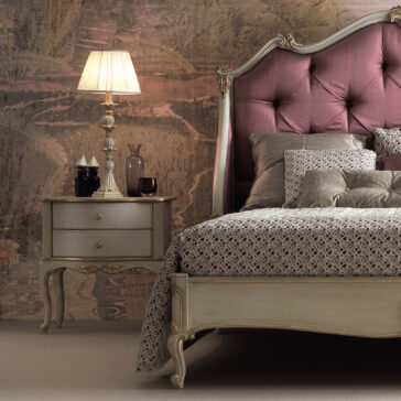 Vintage-style bedroom featuring an Ornate Classic Style Winged Bed with a tufted purple headboard. A winged bedside table with two drawers is adorned with a classic lamp with a fabric shade, decorative jars, and a dark glass vase. The room is tastefully finished with elegant, muted-pattern wallpaper.