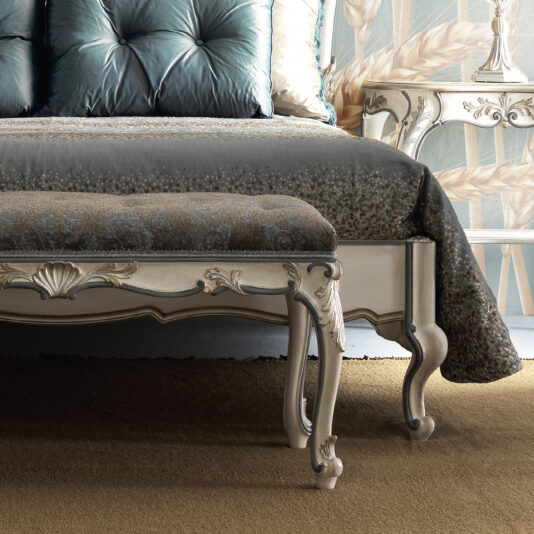 A lavish bedroom arrangement featuring an exquisite tufted headboard, a beautifully patterned duvet, and the Ornate Louis Style Bench with its intricately carved legs. The backdrop displays a floral-designed wallpaper, accentuated by a vintage nightstand adorned with elegant details.