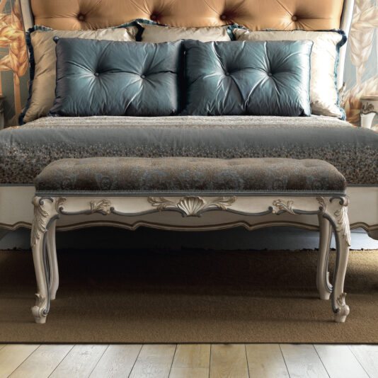A luxurious bedroom showcases a plush bed upholstered in blue, complemented by two large cushions. At the foot of the bed, an Ornate Louis Style Bench, featuring intricate carvings, enhances the setting with its elegance and comfort.
