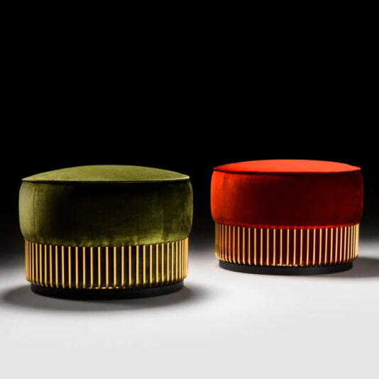 Two round ottomans against a dark background. One is upholstered in green velvet with a gold base, and the other in red velvet with a similar gold base. Both rest on a shadowed surface.