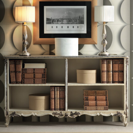 A Large Rococo Inspired Sideboard, adorned with ornate detailing, holds antique books and round beige boxes. Two silver lamps with beige shades sit on top, along with a framed black and white photo of a bridge against the textured wall's circular patterns.