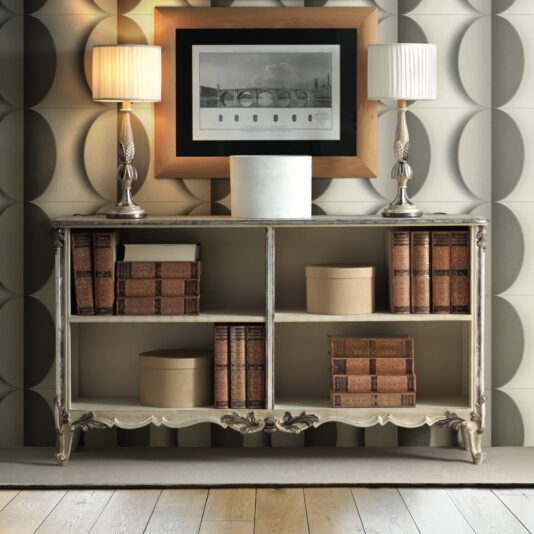 A vintage wooden bookshelf with ornate carvings stands alongside the Large Rococo Inspired Sideboard, holding books and round boxes. Two table lamps with white shades sit on top, flanking a framed black-and-white photo. The wallpaper features a geometric circle pattern.