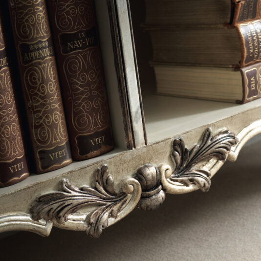 A close-up view of a detailed wooden bookshelf, similar to the Large Rococo Inspired Sideboard (Copy), filled with vintage books featuring embossed leather spines. The bookshelf showcases elaborate carved details, with some books artfully stacked horizontally.