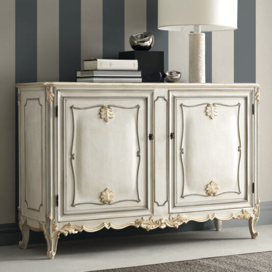 The Rococo Inspired Two Door Sideboard exudes vintage elegance with its ornate gold embellishments. Set against vertical striped wallpaper, the sideboard's two doors feature decorative carved accents. A contemporary lamp and a stack of books add to the sophisticated display.