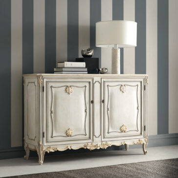 The Rococo Inspired Two Door Sideboard, featuring intricate gold detailing, is positioned against a striped wall. Its top is adorned with a sleek modern white lamp and two small silver sculptures, while a grey carpet peeks into view on the floor.
