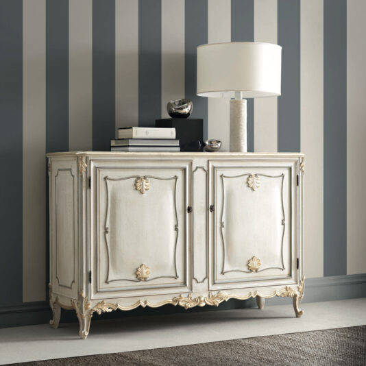 The Rococo Inspired Two Door Sideboard, featuring intricate gold detailing, is positioned against a striped wall. Its top is adorned with a sleek modern white lamp and two small silver sculptures, while a grey carpet peeks into view on the floor.