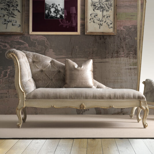 A vintage chaise lounge with tufted upholstery and ornate wood carvings rests on a light wood floor. Behind it, the Rococo Style Wall Mirror complements the floral artwork and decorative wallpaper. A single pillow is placed on the chaise.