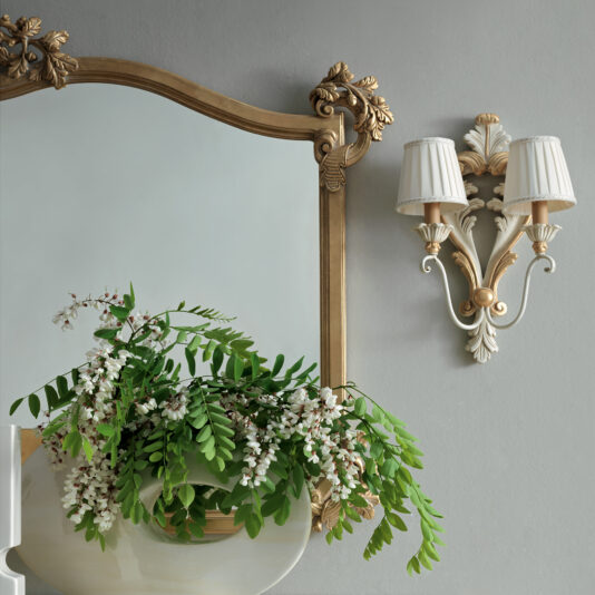 In a decorative setting, an ornate mirror with a golden frame is complemented by the Rococo Style Double Wall Light. Beneath them, there's a white vase filled with green foliage and small white flowers, all set against a soft gray wall.