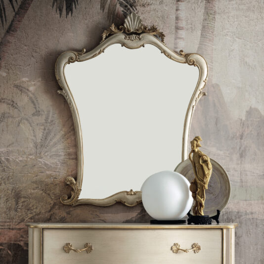 A Rococo-Style-Wall-Mirror-1 graces the wall, reflecting a subtle palm tree mural. Below it, a vintage dresser is adorned with a round frosted glass lamp and a small statue, forming an elegant composition.