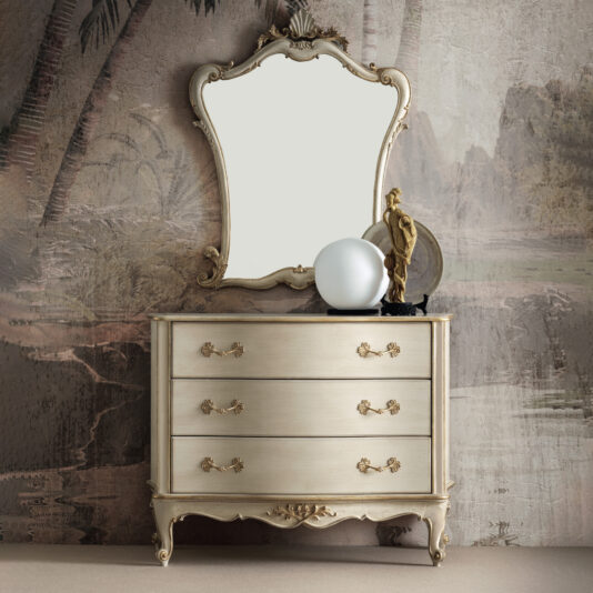 A vintage wooden dresser with ornate gold handles is positioned against a textured, nature-themed mural wall. Placed on top of the dresser are a decorative statue and a spherical frosted lamp. Mounted above is the Classic Rococo Style Overmantel Mirror (Copy), featuring a curved frame that enhances the elegant ambiance.