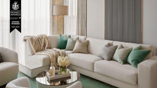 A modern living room with a white sectional sofa adorned with green and grey pillows. A light throw is draped over the corner. A round glass coffee table holds decorative items and flowers. A floor lamp stands in the background. Prestigious UK Property Award winners badge is overlaid