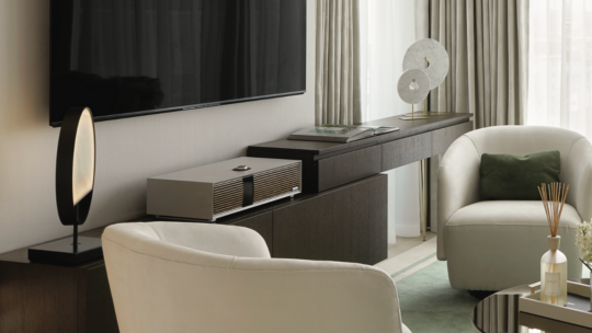 A modern living room, worthy of a UK Property Award, features a large wall-mounted TV, white armchairs, and a sleek sideboard with a sound system. Circular sculptures and a green rug add elegance as soft natural light filters through light-colored curtains.