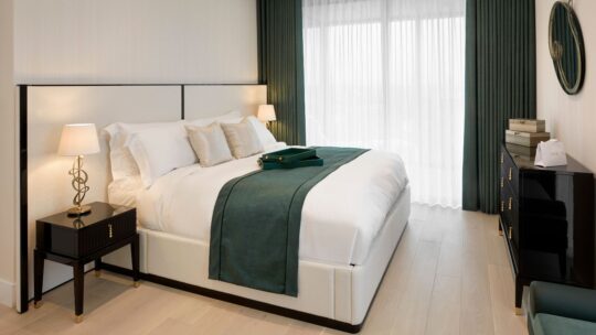 A modern bedroom, deserving of a UK Property Award, showcases a large bed with white and green bedding. It includes two bedside tables with lamps, a dresser, and a circular mirror. Large windows with green curtains flood the room with natural light.