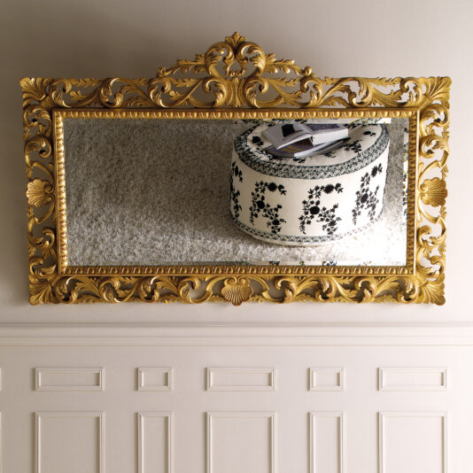 A Baroque Style Overmantel Mirror with a gold frame reflects a round cushioned stool adorned with black floral patterns. The ornate mirror captures the image of a white carpet and several books resting on the stool, while the wall beneath is elegantly decorated with white paneling.