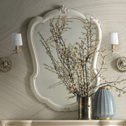 A decorative wall features the Curved Rococo Style Mirror, showcasing an ornate frame. In front, a metal vase cradles branches adorned with small white flowers. Elegantly mounted on either side are two wall sconces with white shades, enhancing the vintage charm of the setup.