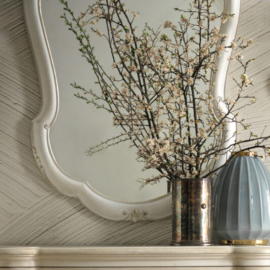 Curved Rococo Style Mirror A decorative vignette showcases the Curved Rococo Style Mirror alongside a ceramic vase filled with small blossom branches, contrasting beautifully against textured wallpaper. This arrangement is set on a light-colored surface, crafting an elegant and serene atmosphere.