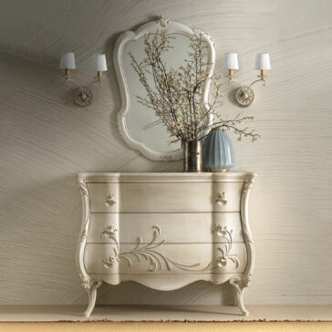 Curved Rococo Style Mirror Against a textured beige wall stands a vintage white dresser featuring ornate floral carvings. Above the dresser hangs the Curved Rococo Style Mirror, framed by elegant wall sconces. Atop the dresser, you'll find a vase with branches and a blue glass lamp.