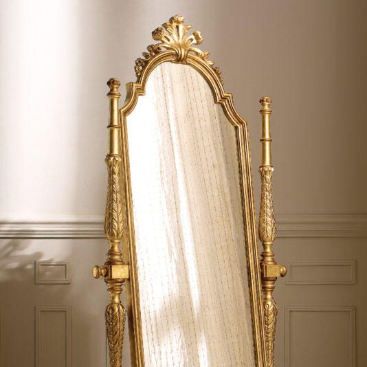 Freestanding Rococo Style Tilting Mirror A Rococo-style freestanding mirror with a gold frame, known as the Freestanding Rococo Style Tilting Mirror, stands against a cream-colored wall. The intricate decorative details at its top enhance its elegance, and the tilting feature allows it to reflect soft natural light while casting gentle shadows on the wall beside it.
