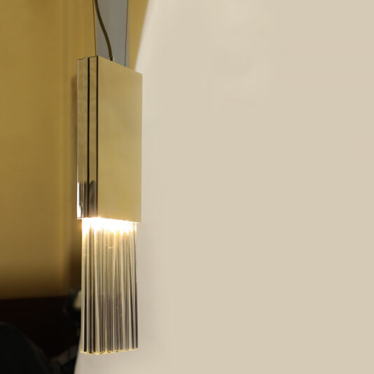 A modern wall lamp with a sleek, gold rectangular design boasts a fringe of vertical metal rods similar to the Glass Drop Pendant Light at the bottom, casting a warm glow against a light-colored background.
