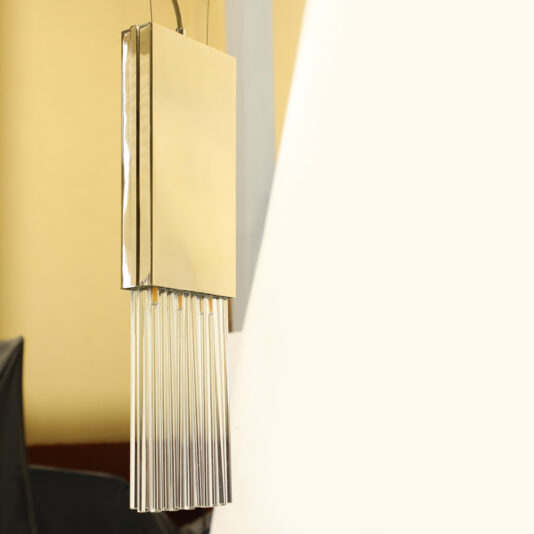 The stylish Glass Drop Pendant Light features vertical glass rods that gracefully hang from a rectangular gold fixture, beautifully contrasted against a gentle, light backdrop.