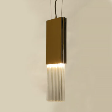 Slim Glass Drop Pendant Light The Glass Drop Pendant Light features a contemporary design with a rectangular golden metal top and vertical glass rods below, casting a warm glow against a simple beige backdrop.