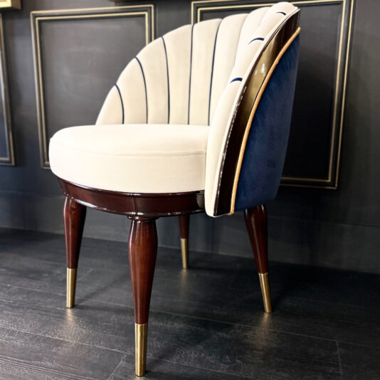 The High End Art Deco Style Occasional Chair boasts a chic white upholstered seat and backrest, accented with dark blue tones and gold trim. This elegant piece is complemented by dark wooden legs capped with gold, making it a perfect match for any dark paneled wall.