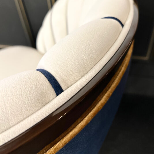 Close-up of the Art Deco Style Occasional Chair featuring white tufted cushions and navy blue fabric. The brown trim offers a striking contrast, enhancing its sophisticated design, while the dark background highlights the chair's elegance.