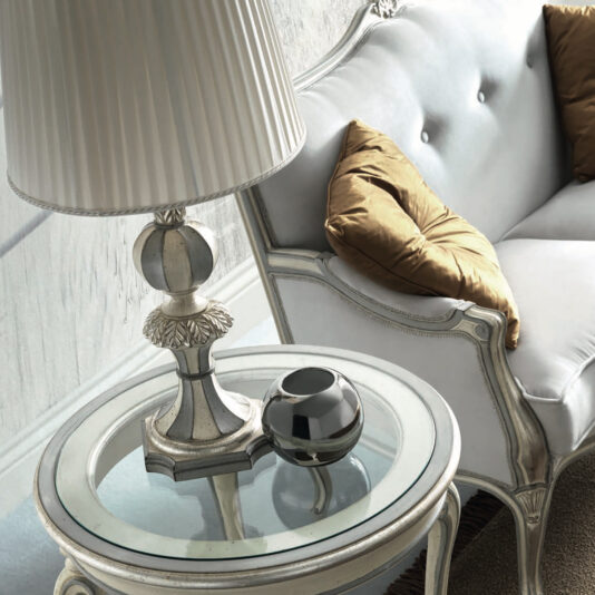 A sophisticated living room scene features the High End Rococo Style Sofa, a white tufted masterpiece embellished with golden cushions. It is complemented by a round glass-top side table and a decorative silver lamp, with a small black and silver ornament placed beside it to complete the elegant setting.