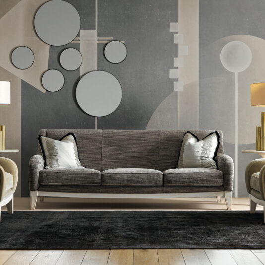 Large Art Deco Style Sofa A contemporary living room featuring the Large Art Deco Style Sofa in gray, accentuated with two pillows, and complemented by table lamps on either side. The wall showcases a geometric design adorned with circular mirrors, and a black rug embellishes the wooden floor.
