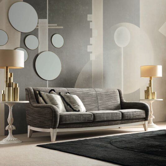 Large Art Deco Style Sofa A modern living room is highlighted by a Large Art Deco Style Sofa in gray accompanied by two white side tables, each adorned with a gold lamp. The wall is decorated with abstract geometric shapes and round mirrors, while a black area rug beautifully enhances the light gray and cream color scheme.