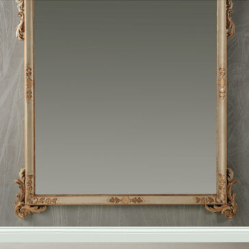Large Baroque Style Wall Mirror The Large Baroque Style Wall Mirror, featuring an intricately carved wooden frame, is mounted on a wall adorned with patterned wallpaper; its ornate design captures the timeless elegance of bygone eras. The mirror reflects the texture of the wall along with a portion of the floor beneath.