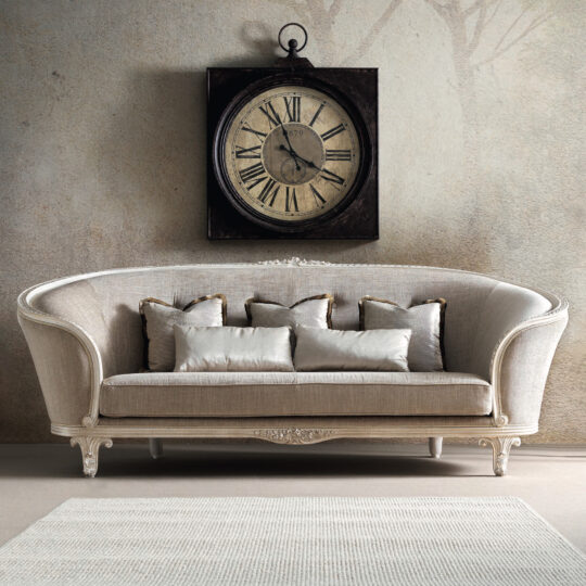 A vintage-inspired room is centered around the Large Rococo Style Curved Sofa, elegantly upholstered in beige and adorned with five plush cushions. Above the sofa, an antique-style clock with Roman numerals adds to the cozy and sophisticated atmosphere, complemented by a neutral-toned rug on the floor.