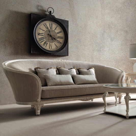 Large Rococo Style Curved Sofa A stylish living room showcases a vintage-style clock with Roman numerals on the wall above the Large Rococo Style Curved Sofa, which is adorned with multiple cushions. In front of the sofa sits an elegant round glass coffee table with an ornate base.