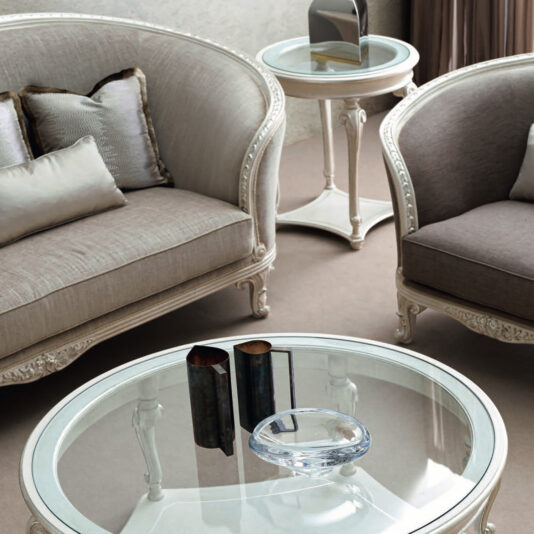 Large Rococo Style Curved Sofa In the stylish living room, the centerpiece is a Large Rococo Style Curved Sofa paired with another upholstered seat, all arranged around a circular glass coffee table. This table features a decorative bowl and two cylindrical vases. In the backdrop is a coordinating side table with a curvilinear design in neutral shades.