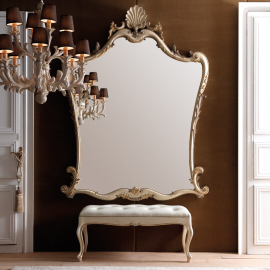 The elegant room features a Large Rococo Style Wall Mirror with a decorative frame. Below the mirror is a cushioned bench with elegantly curved legs. On the left, a classic chandelier adorned with black lampshades hangs, offering a striking contrast to the brown and white walls.
